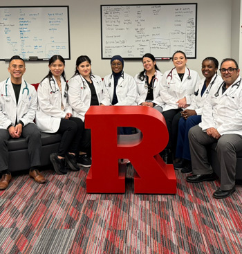 8 students in lab coats sitting on a grey couch.  There is a Rutgers Block R sculpture in front, and a whiteboard in the back.