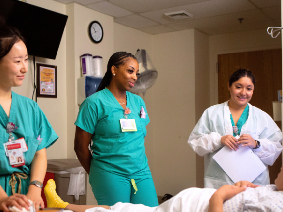 3 Midwifery students at bedside.  Two are in TEal scrubs, one has a white coat