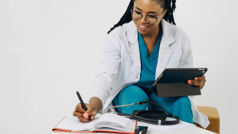 Female nurse wearing glasses and wearing a white coat and scrubs holding a tablet while writing into an orange book. There is a stethoscope on the table.